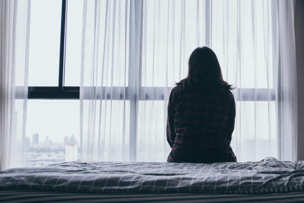 a person sits on a plain bed looking out a window while experiencing benzodiazepine abuse