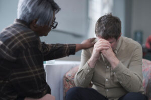a patient struggles with the long term effects of trauma and puts his head to his hands appearing distraught while his therapist consoles hime by resting his hand on the patients shoulder