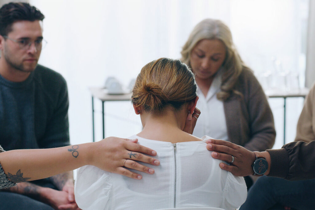 a woman participates in support groups for addiction and has two group members console her by patting each of her shoulder