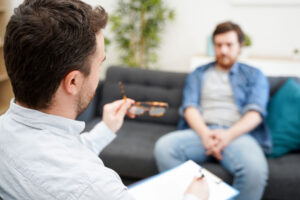 a therapist with his notepad in his lap explains DBT for anxiety to a patient who sits on a couch across from him