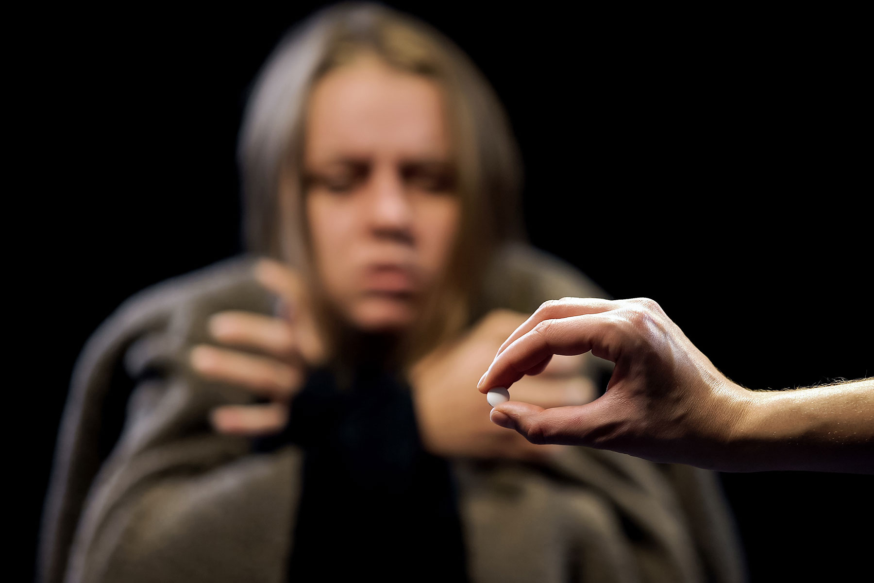 a person shivers in the background in a blanket while someone in the foreground holds up a pill to show painkiller abuse