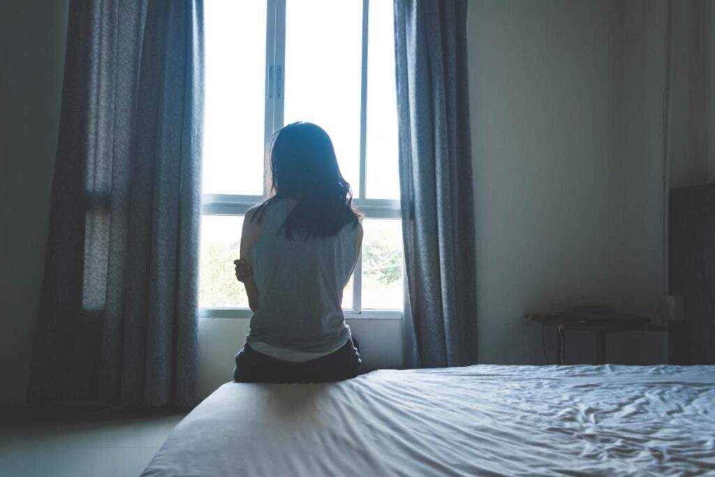 a person sits on the edge of a bed looking out a window to show trauma and alcohol abuse